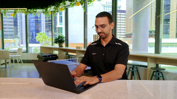 Man wearing smart casual work attire in an office sitting at a bench using a laptop.
