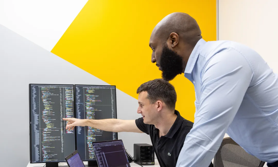 Two IT professionals at a desk inspecting multiple computer and laptop screens that display code