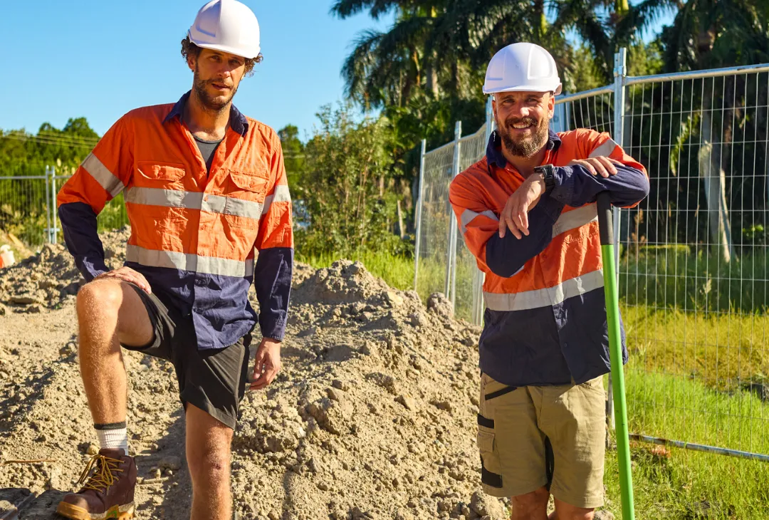 Two landscapers wearing safety gear standing at a worksite with sand and tools.