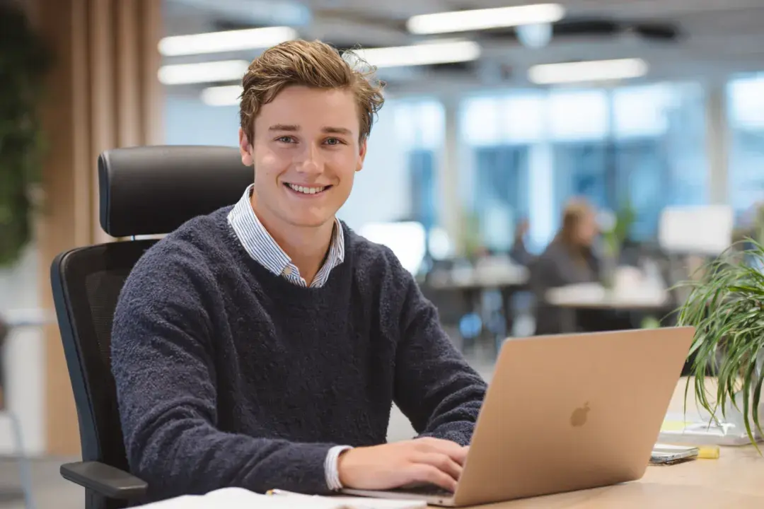 Man wearing a sweater and button shirt sitting at a desk with a laptop smiling