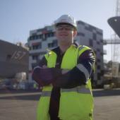 A person in a ship yard wearing a trades uniform with his arms crossed and smiling