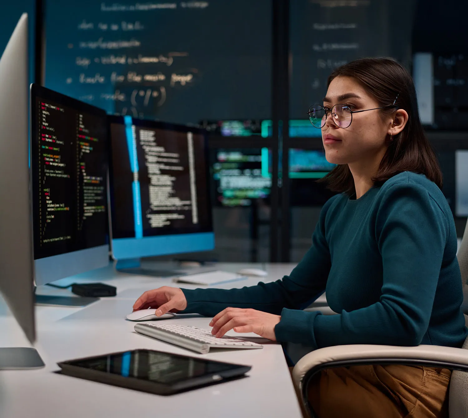 Woman wearing glasses and a sweater at a desk on a computer with two screens.