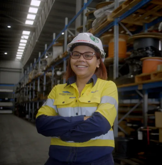 Worker in safety gear smiling in a warehouse with shelves of equipment behind.