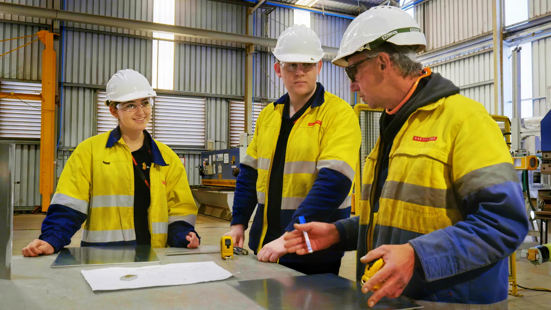Three workers wearing yellow high visibility jackets and protective gear around a work table