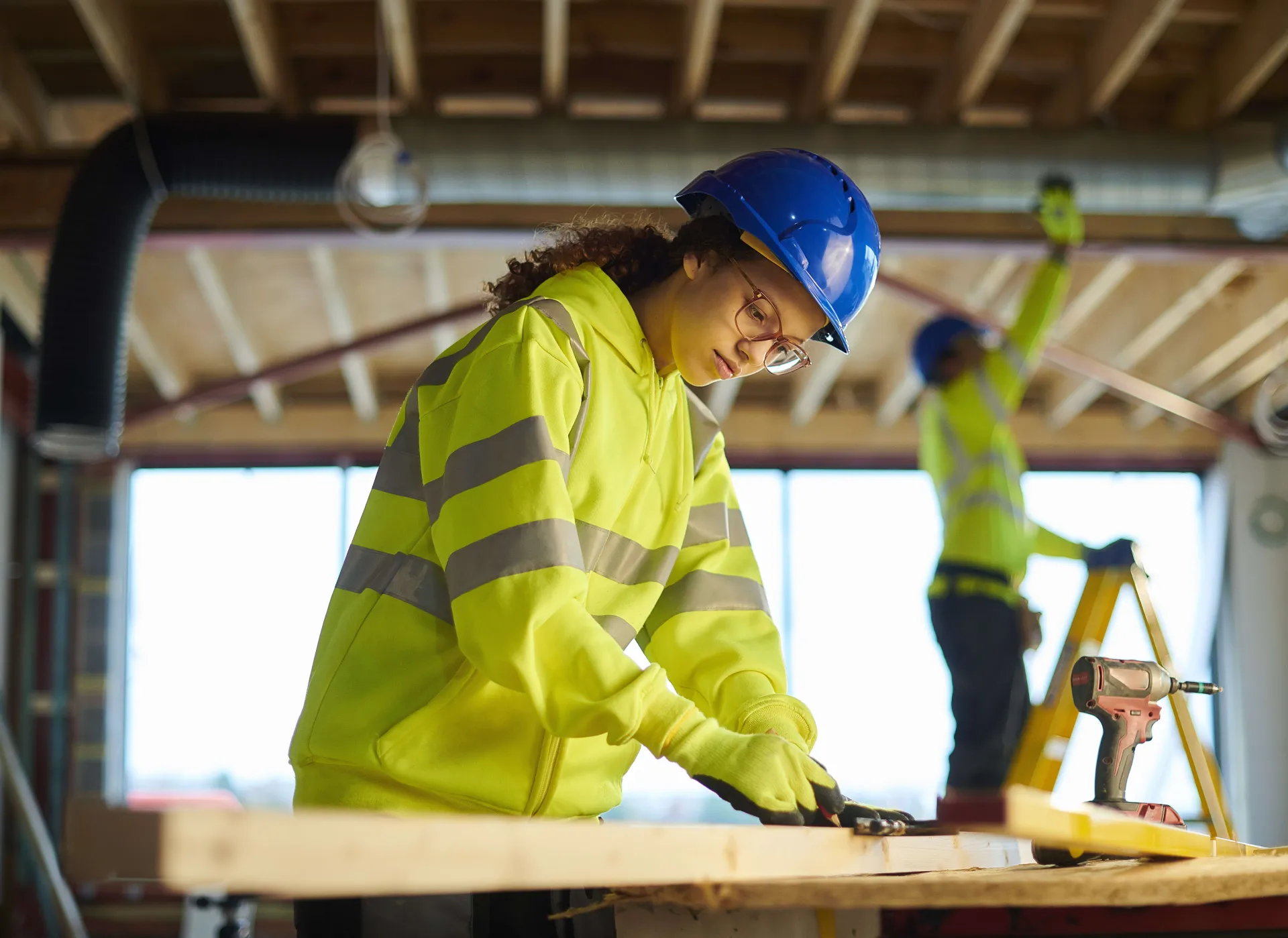 Construction workers in safety gear, one cutting wood and another on a ladder.