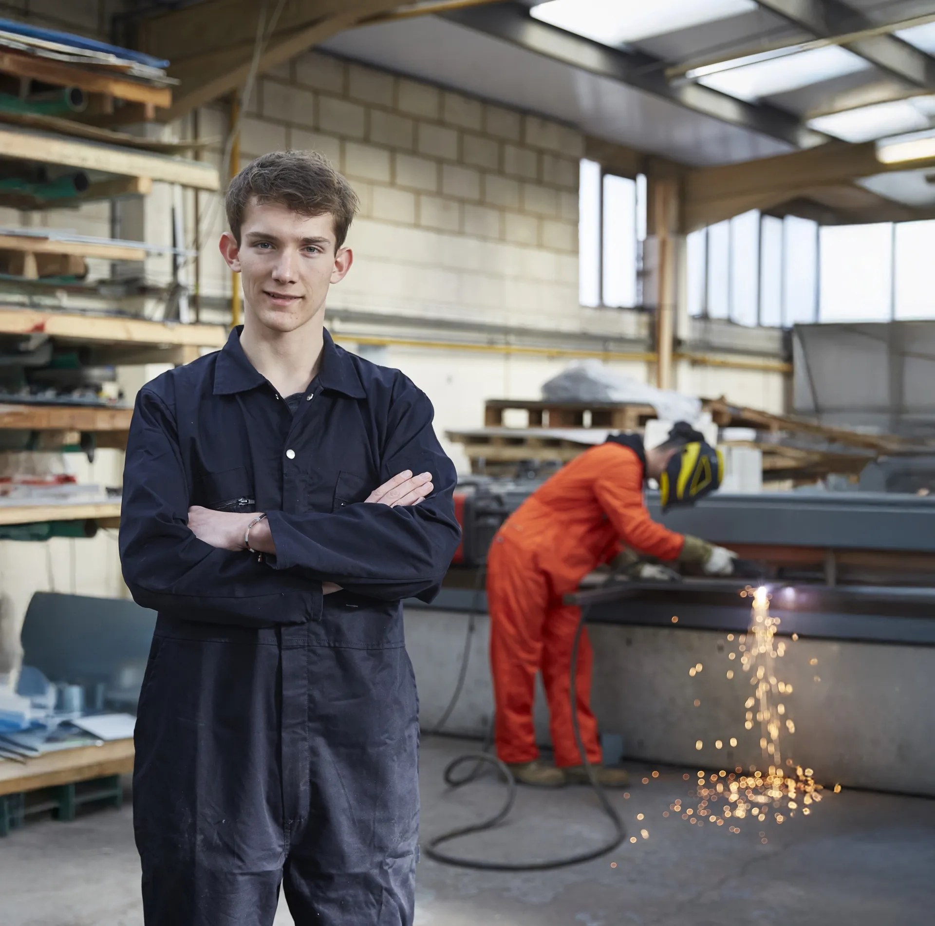 Young worker standing in a warehouse wearing coveralls with his arms crossed.