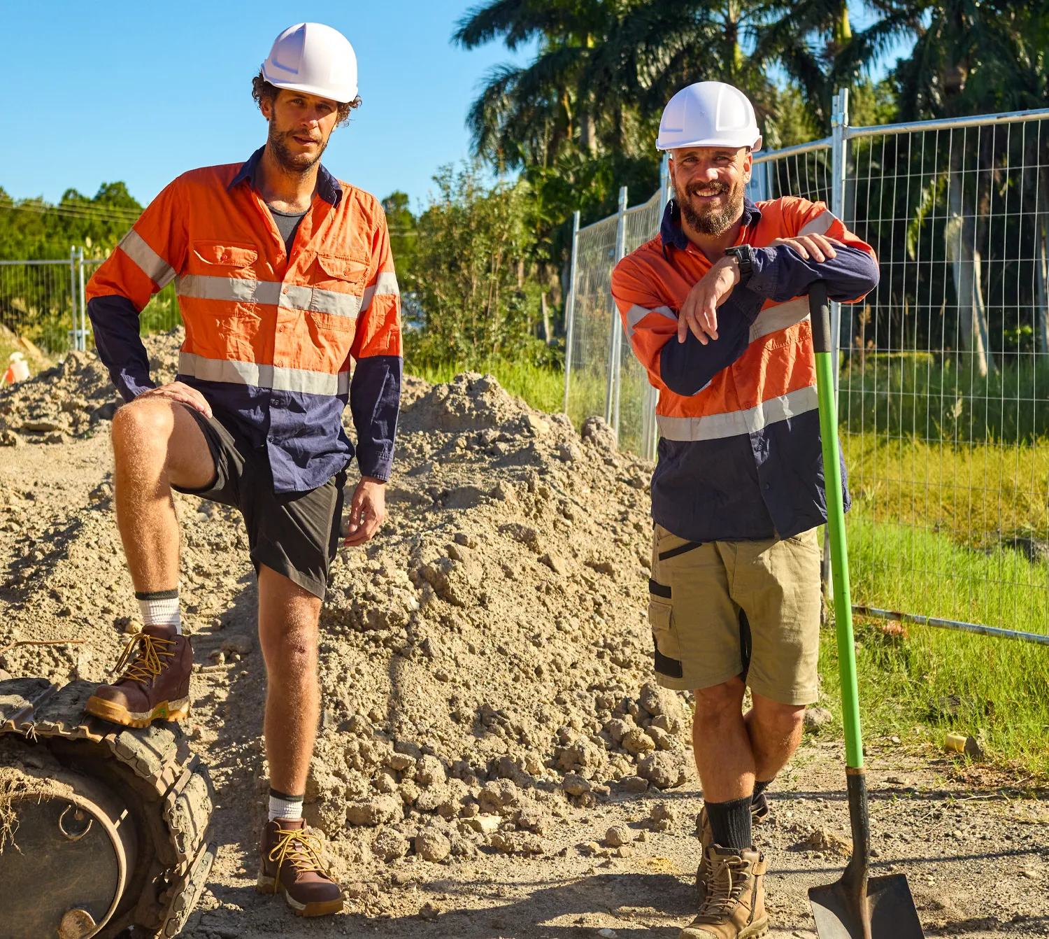 Two landscapers wearing safety gear standing at a worksite with sand and tools.
