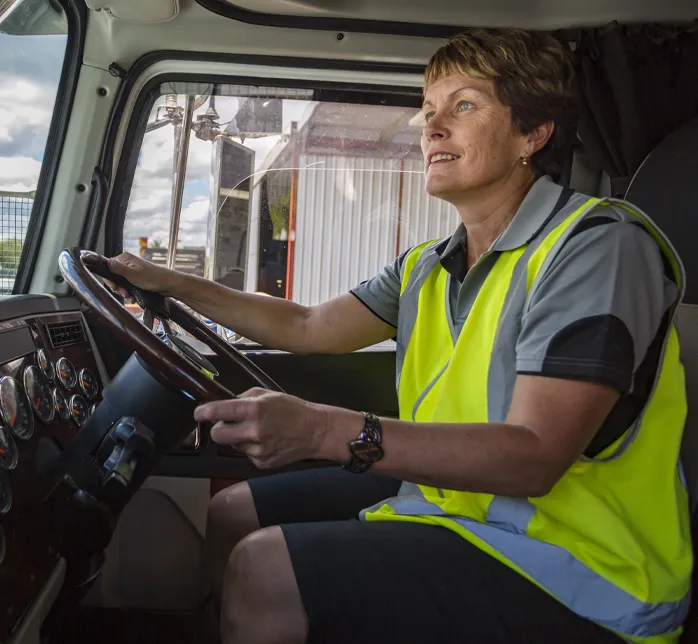 A person wearing a high‑visibility vest sits in the driver’s seat of a large truck, holding the steering wheel and looking ahead with focus.