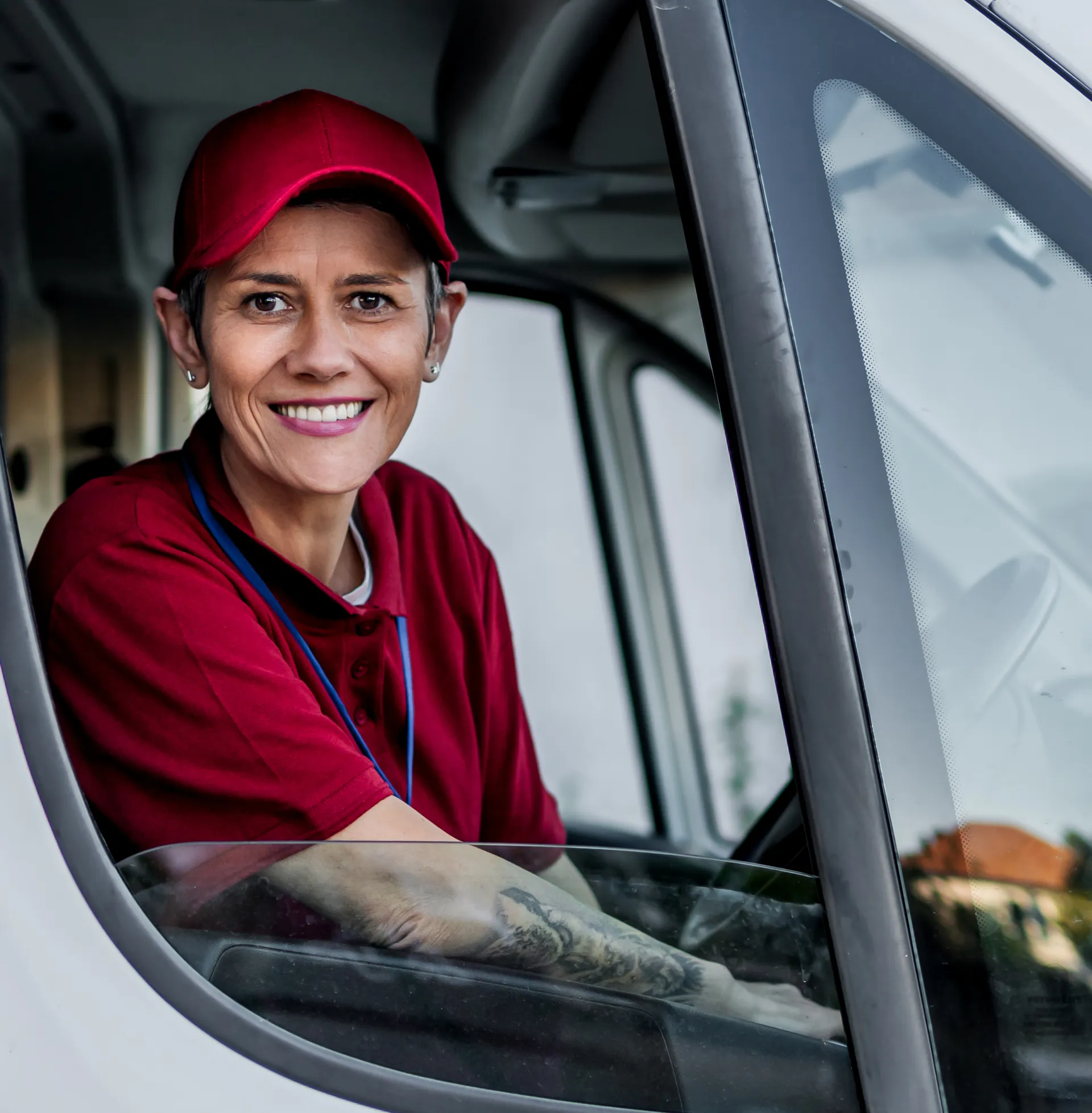 A delivery driver wearing a red uniform and cap smiles while sitting in the driver’s seat of a vehicle, looking out the open window.