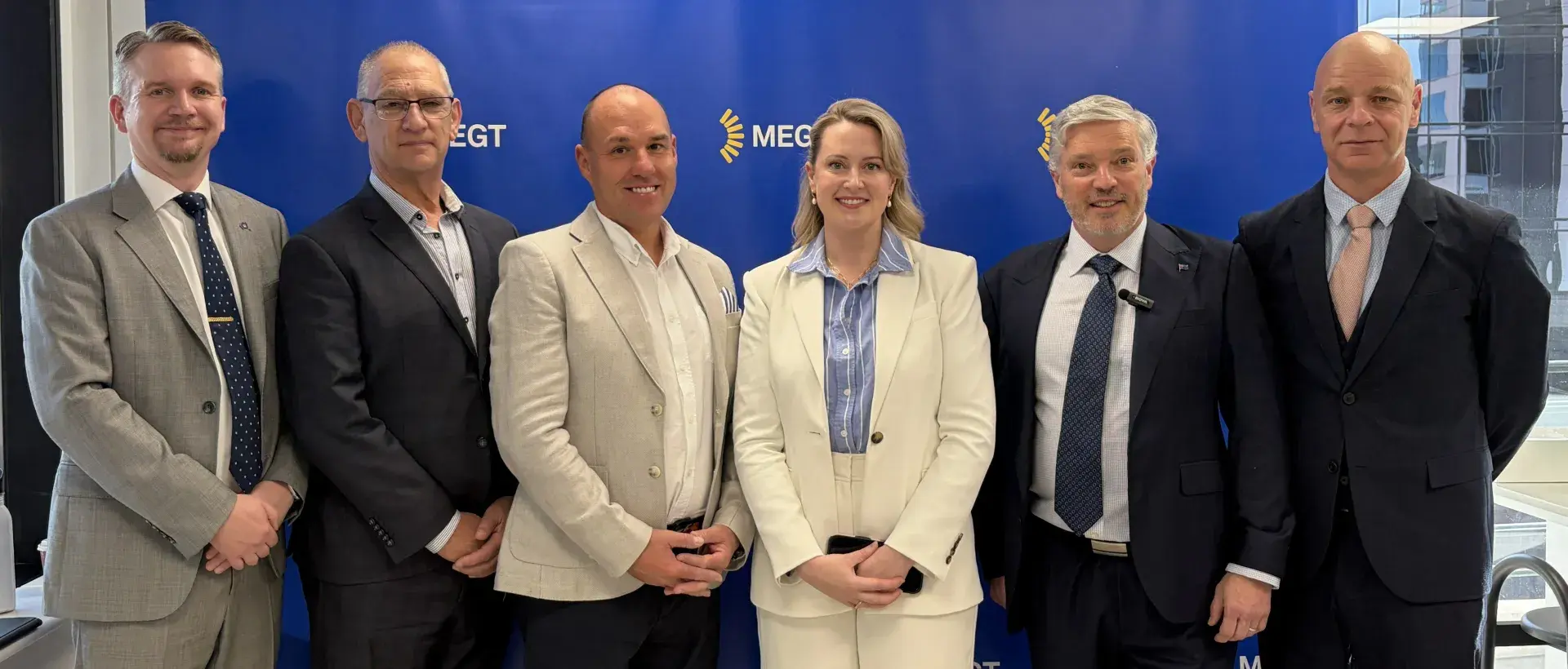 Six people in business attire standing before blue MEGT branded backdrop.