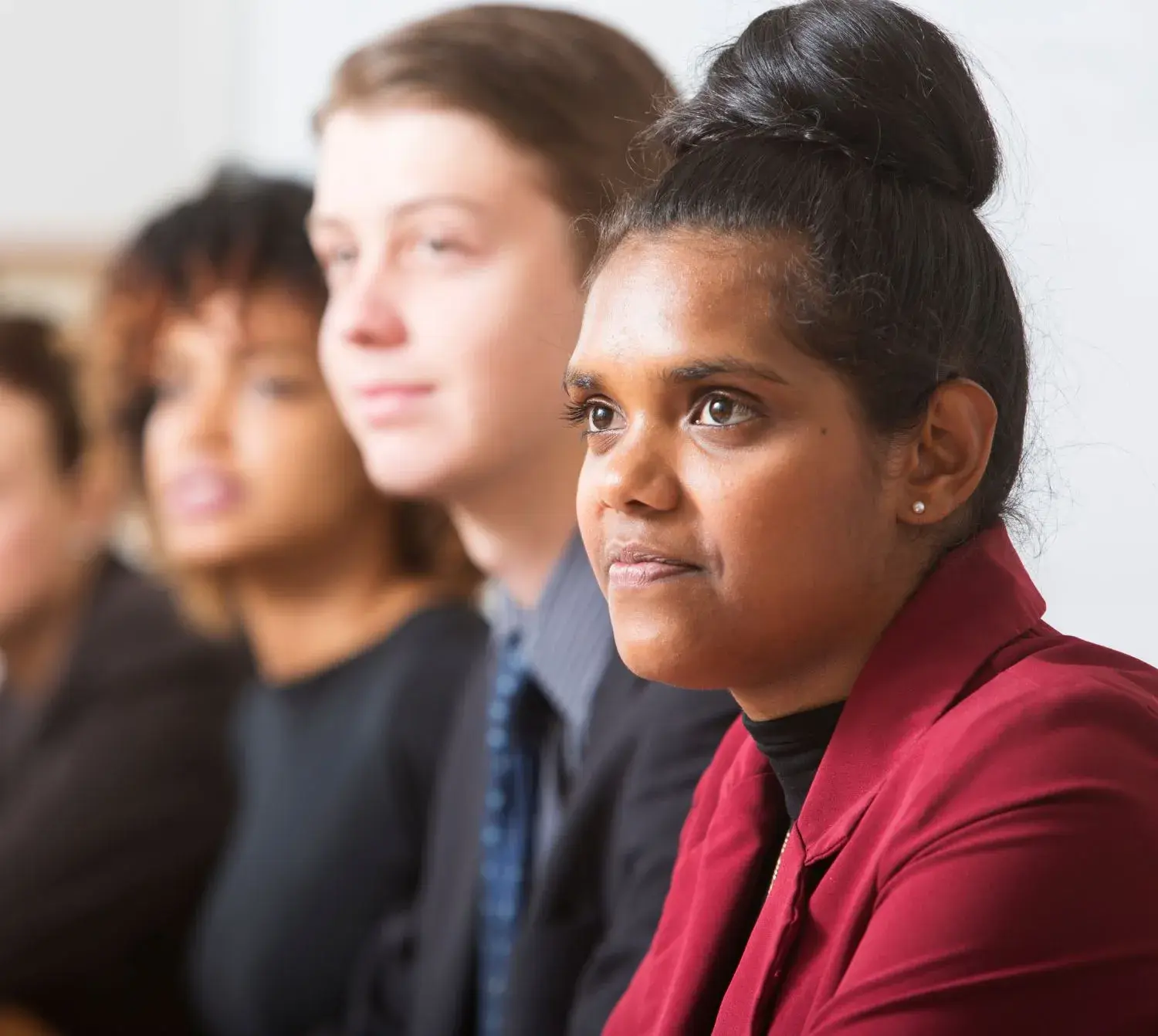 People seated in a meeting, with a person in a red blazer in sharp focus and others blurred in the background.
